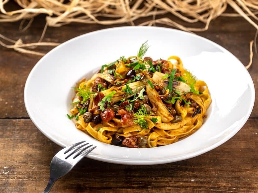 Farfurie cu tagliatelle gatite cu sos rosu, fenicul masline si capere, pe masa de lemn