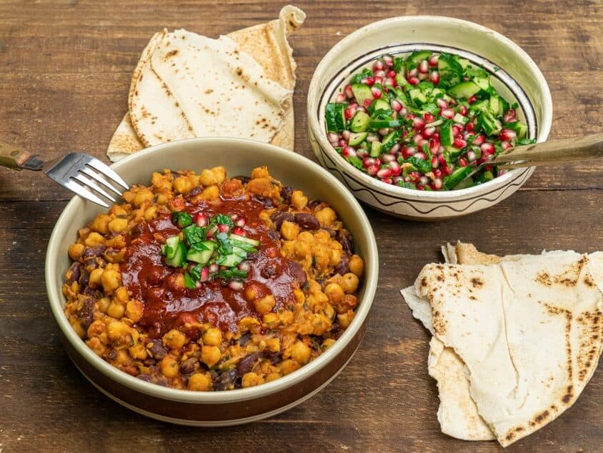 Bol de mancare de naut africana cu topping de pastă harissa, bol cu salsa de rodie si lipii rupte puse pe blat de lemn